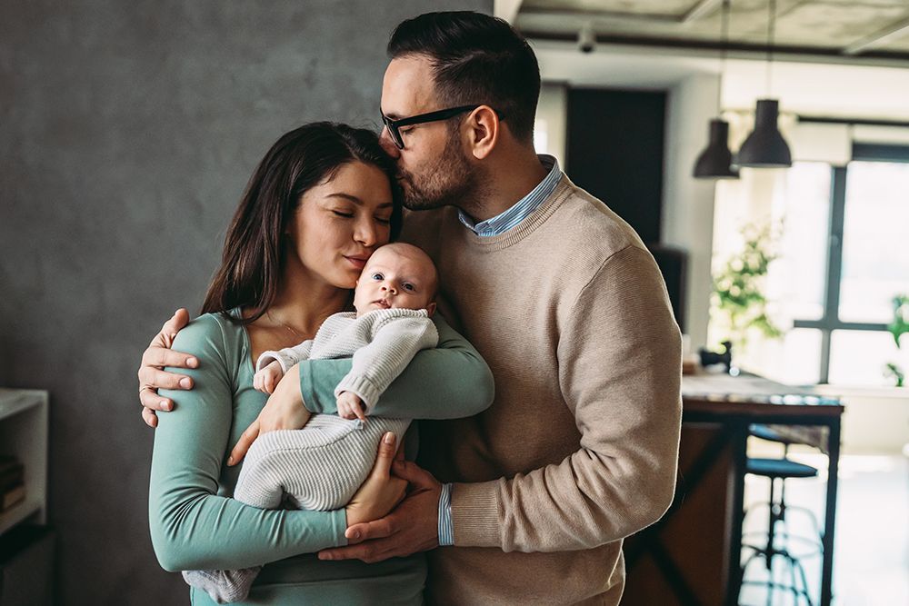 Parents lovingly holding a newborn baby indoors; father kisses mother's forehead; bright, casual interior.