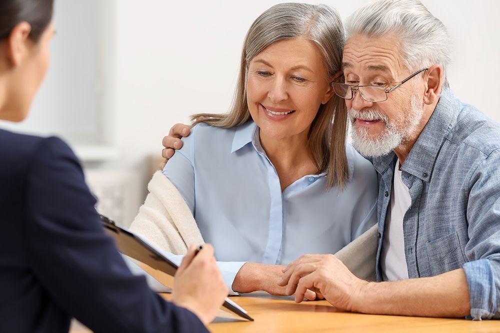 A financial advisor shows paperwork to an older couple. The woman smiles while the man, wearing glasses, looks intently at the document.
