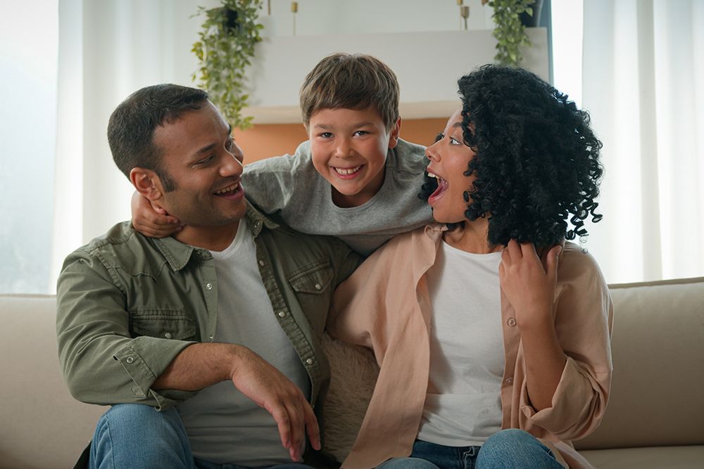 A smiling family of three: a Black father, a biracial child, and a Black mother sitting on a couch indoors.