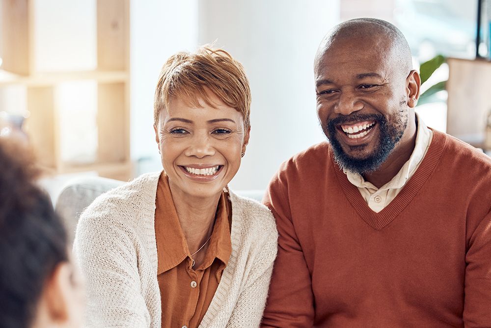 Smiling couple, a Black man and woman, seated in a well-lit room, possibly an office or counseling setting.