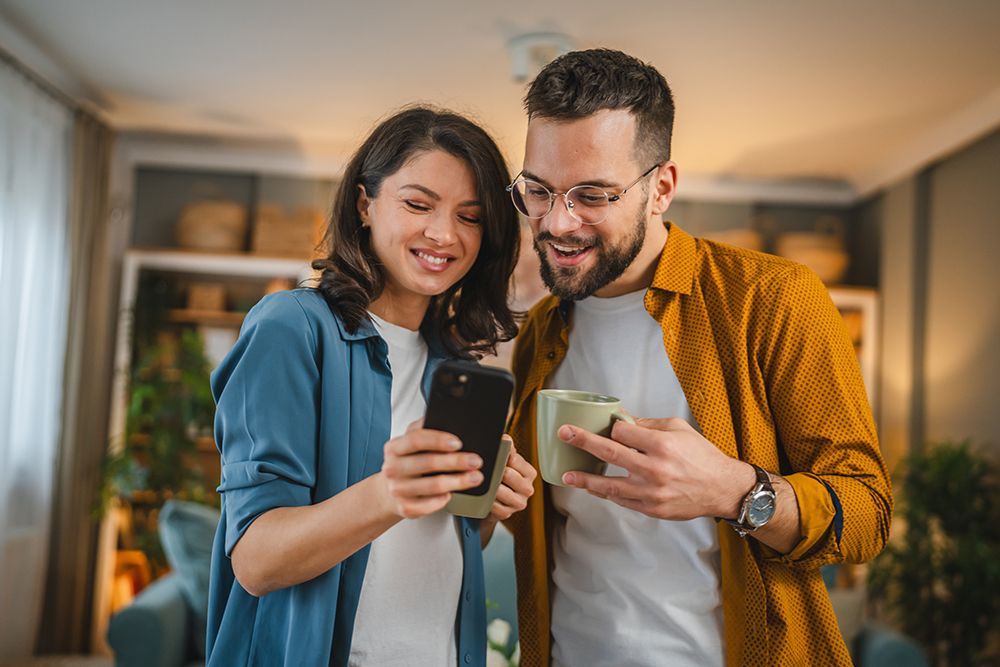 A smiling couple indoors looking at a phone together. The woman holds the phone, and the man holds a mug.