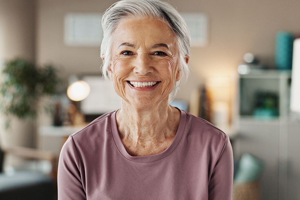 Smiling elderly woman with gray hair, wearing a purple shirt, in a well-lit room.