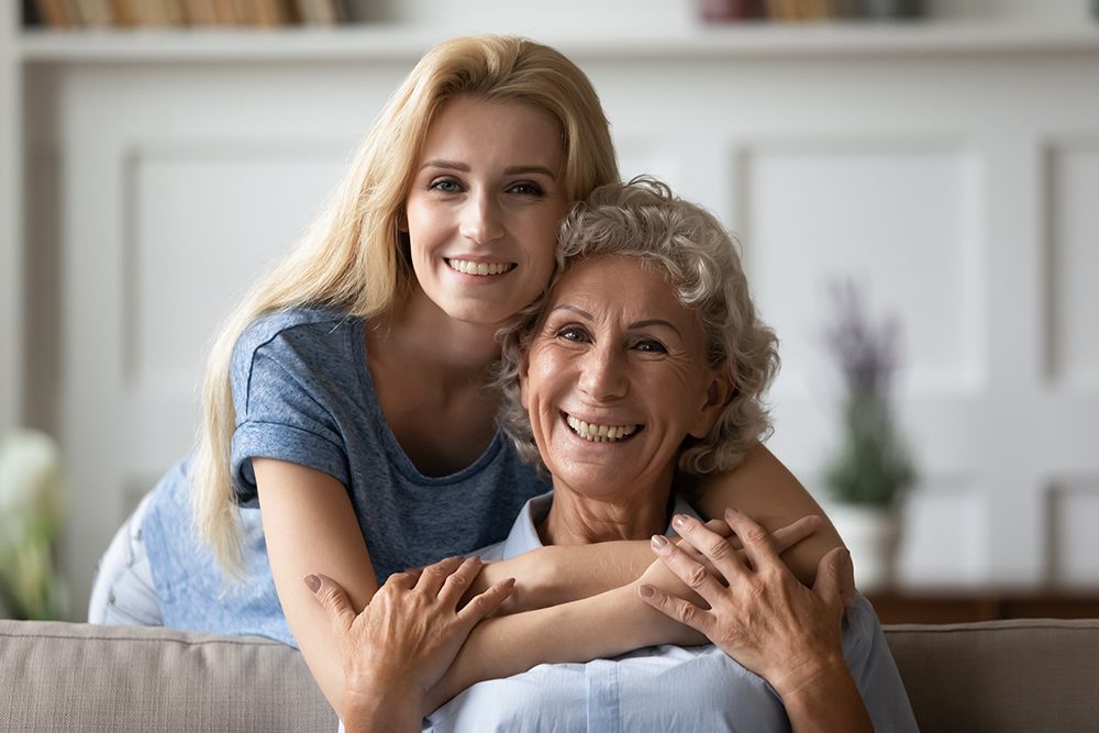 A young woman hugs an older woman, both smiling at the camera. They sit on a couch indoors.