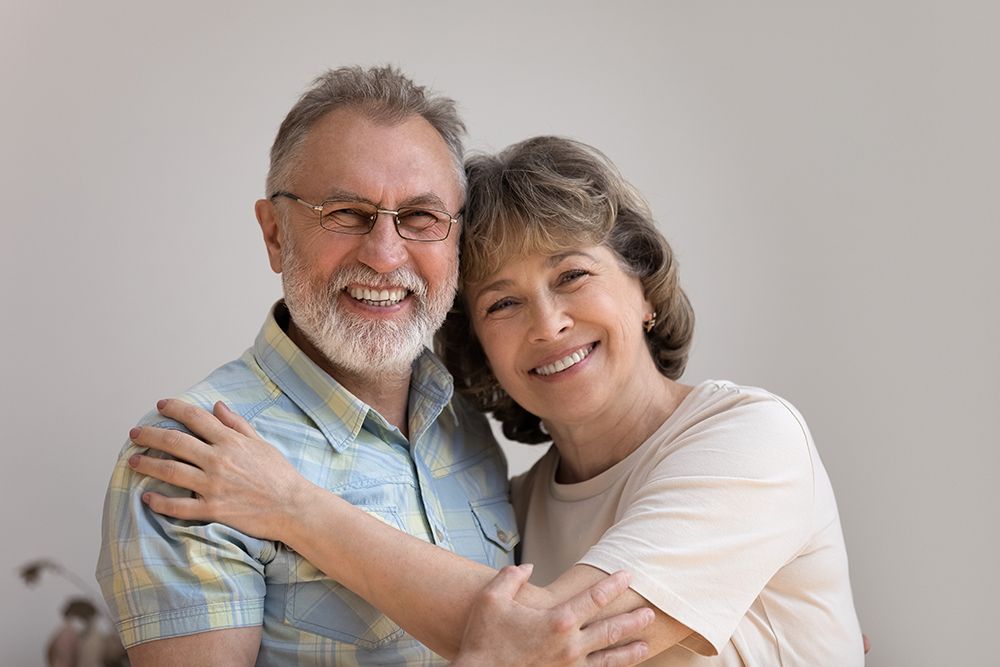 Smiling older couple embracing, indoors; man with glasses and gray beard, woman with short brown hair.