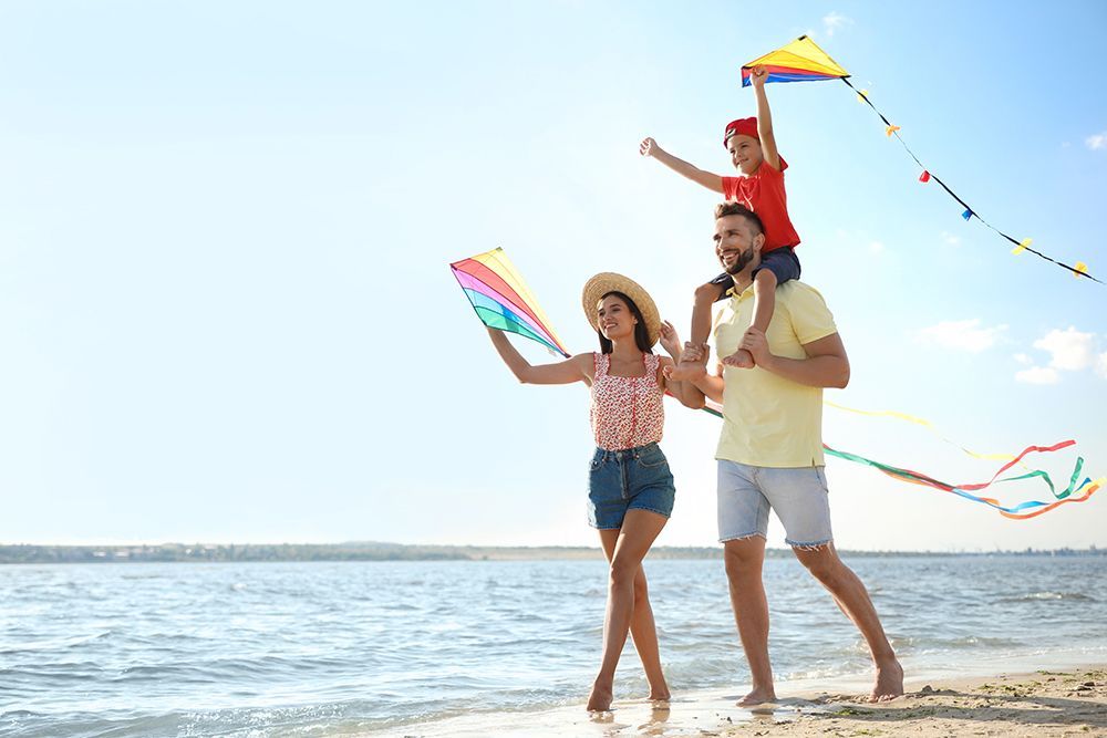 Family flying kites on a sunny beach: a woman, man with a child on his shoulders, holding kites.