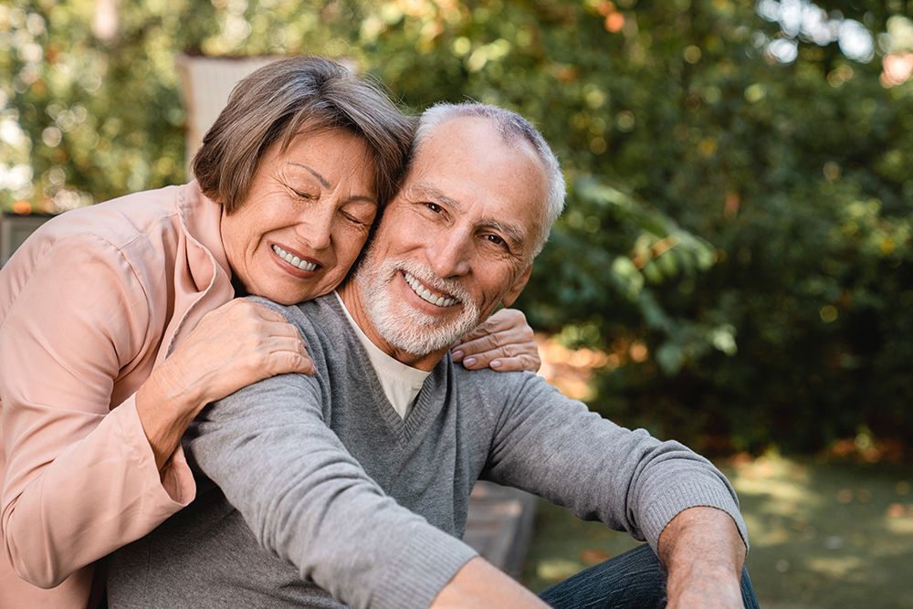 An elderly couple is smiling and embracing outdoors. The woman has her arms around the man's shoulders. They are in a garden.