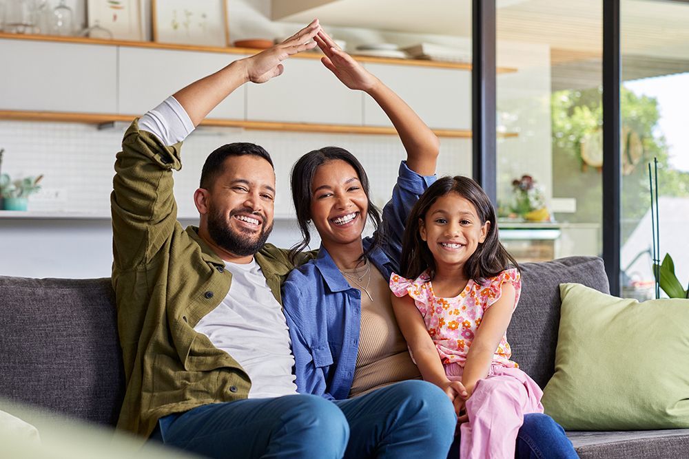 Family of three on a couch, parents forming a roof over their daughter, all smiling indoors.