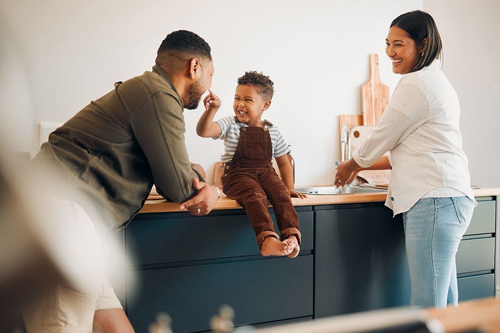 Family of three in a kitchen: a father and son are fist bumping, while the mother smiles, near a countertop.