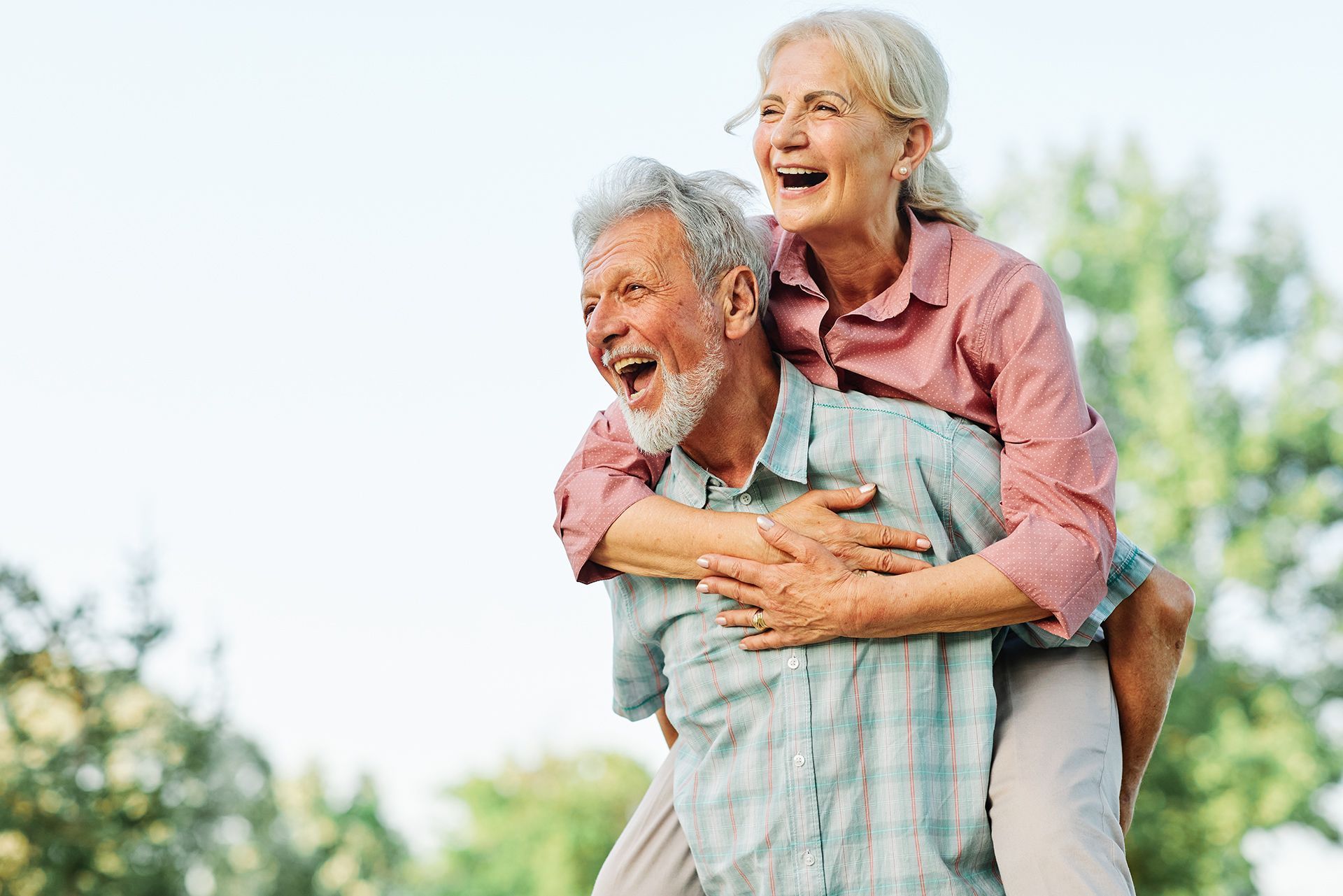 Elderly couple enjoying a piggyback ride outdoors, laughing. Man carrying woman, both smiling in a sunny setting.