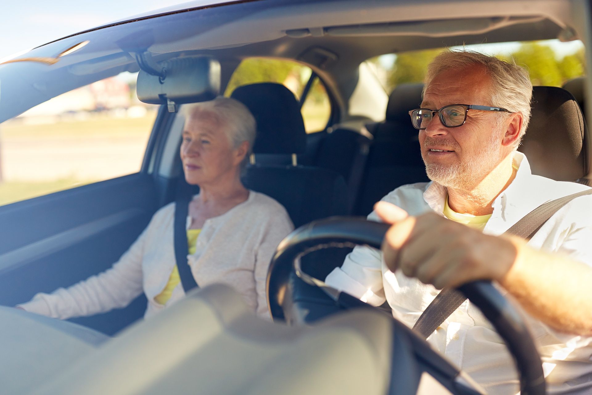 An older man with glasses drives a car, with an older woman in the passenger seat. Both are wearing seatbelts.