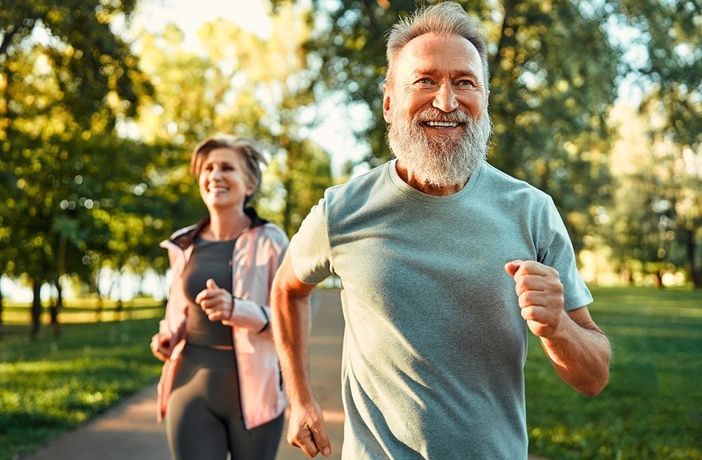 An older man and woman jogging happily on a path in a park on a sunny day. The man has a beard and the woman is smiling.