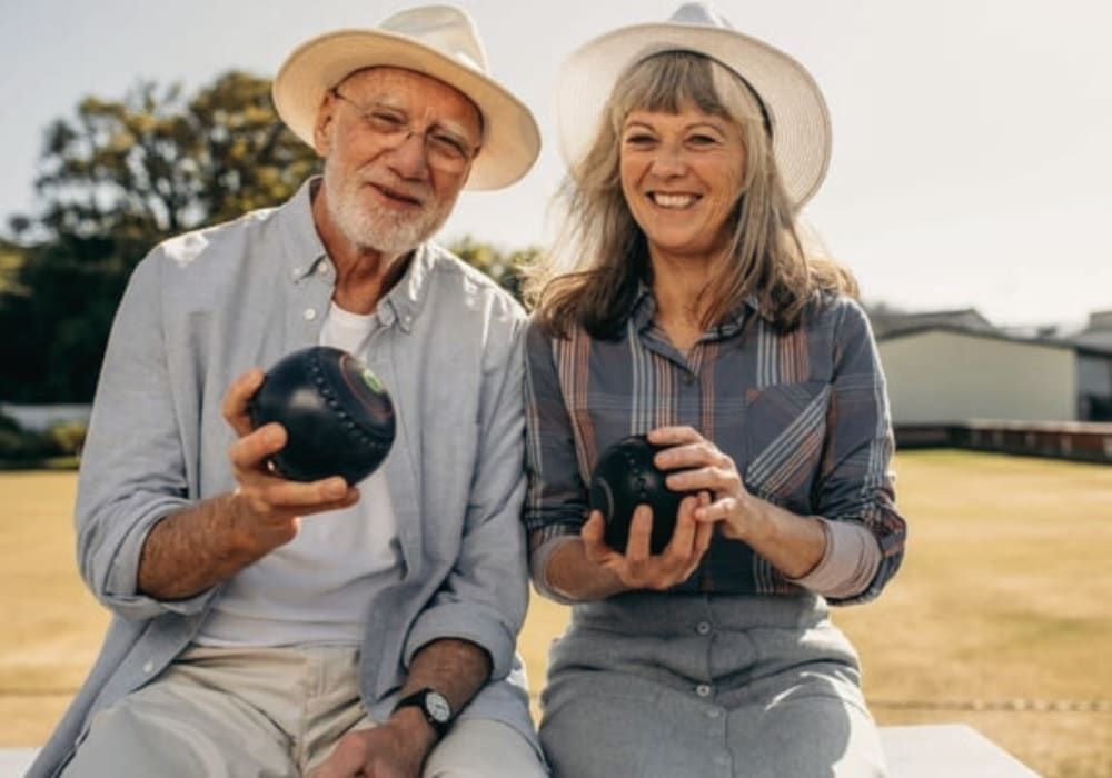Elderly couple smiling, each holding a dark bowling ball, wearing hats, outside.