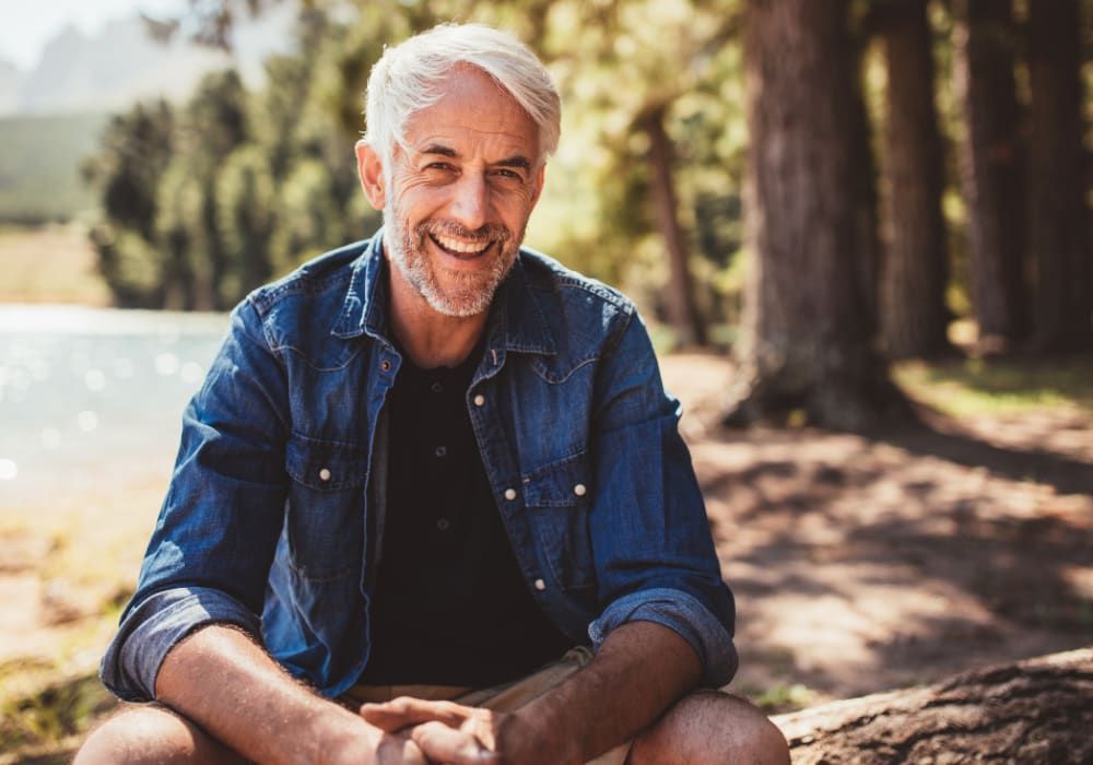 Smiling, gray-haired man in a blue denim shirt sits outdoors by lake and trees.