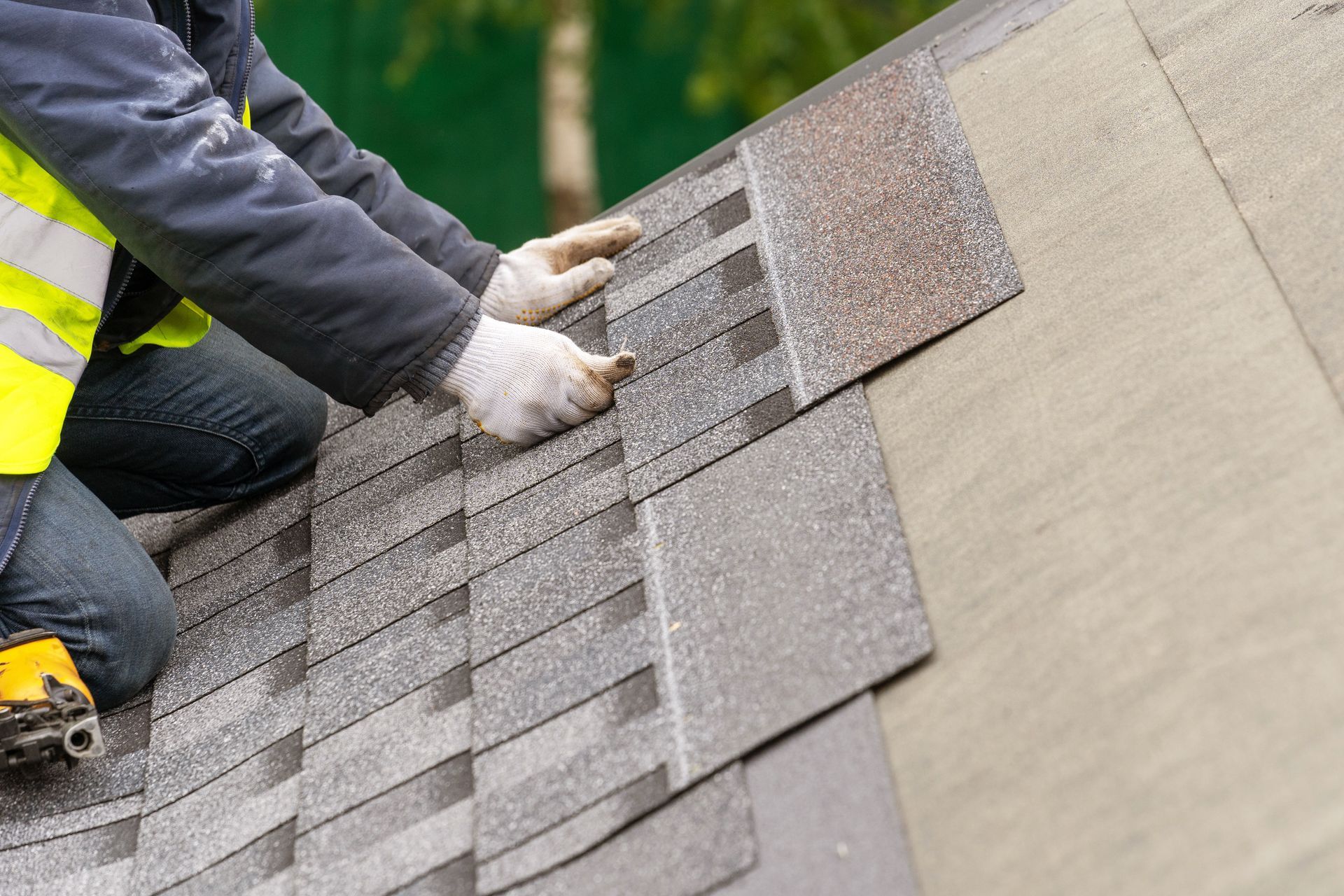 Worker installing asphalt shingles on a roof while wearing gloves and safety gear.