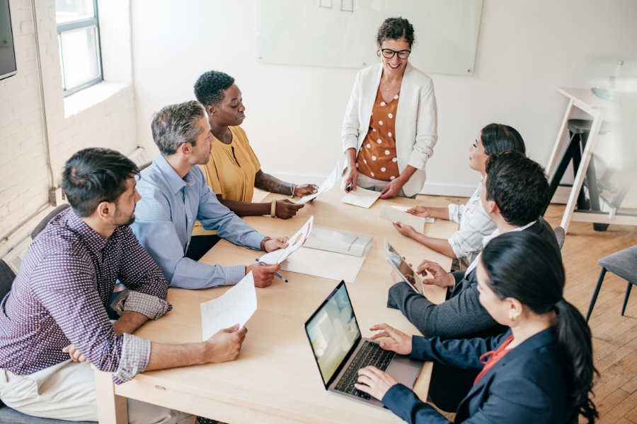 Team meeting around a table, with a presenter standing and colleagues reviewing documents and a laptop.