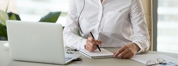 Person in white shirt writing in a notebook beside a laptop at a desk near a window