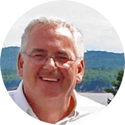 Smiling man in glasses and a white shirt outdoors with water and green hills behind him