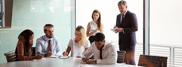Business meeting in a bright office, with people seated around a table and two standing nearby.