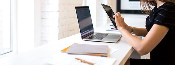Person holding a tablet beside a laptop at a bright desk, with a notebook and pen nearby.