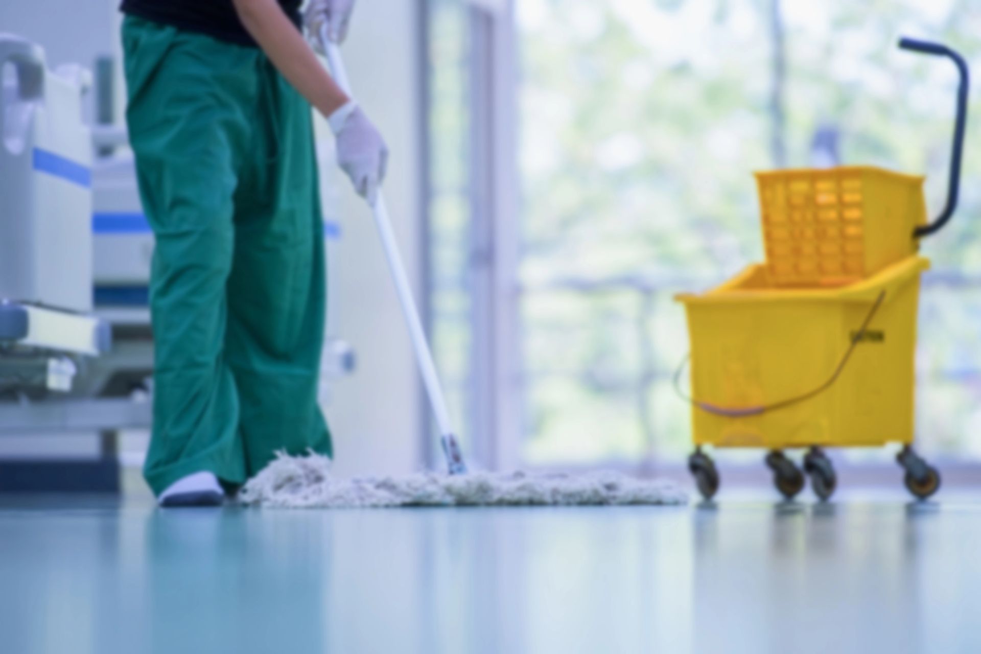 Una persona in camice verde lava il pavimento accanto a un secchio giallo in una struttura medica.