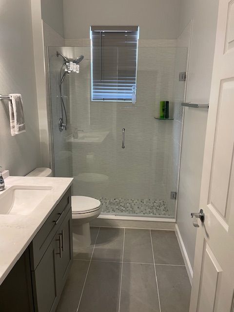 Bathroom with glass shower, white toilet, gray vanity, and window with blinds.