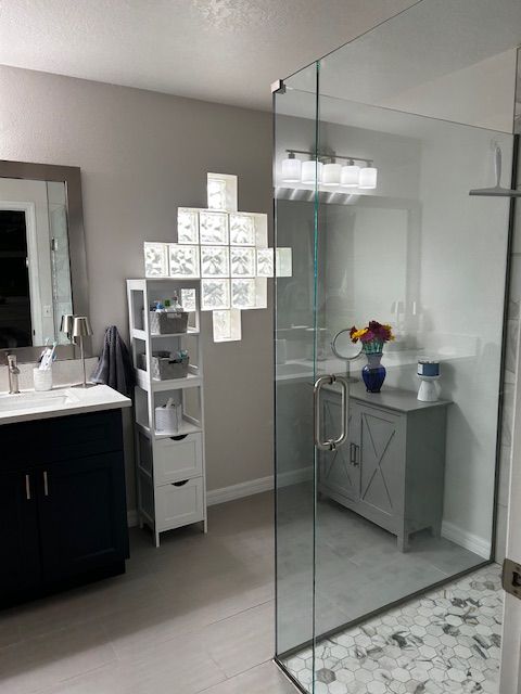 Modern bathroom: glass shower, gray walls, dark vanity, light gray floors, decorative glass block art, and a gray cabinet.
