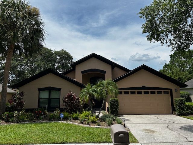 Tan house with black roof, arched entryway, tan garage door, surrounded by palm trees and lush landscaping.