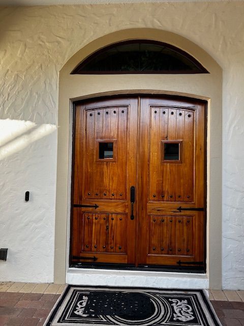 Wooden double doors with decorative metal hardware and two small square windows, under an arched entryway.