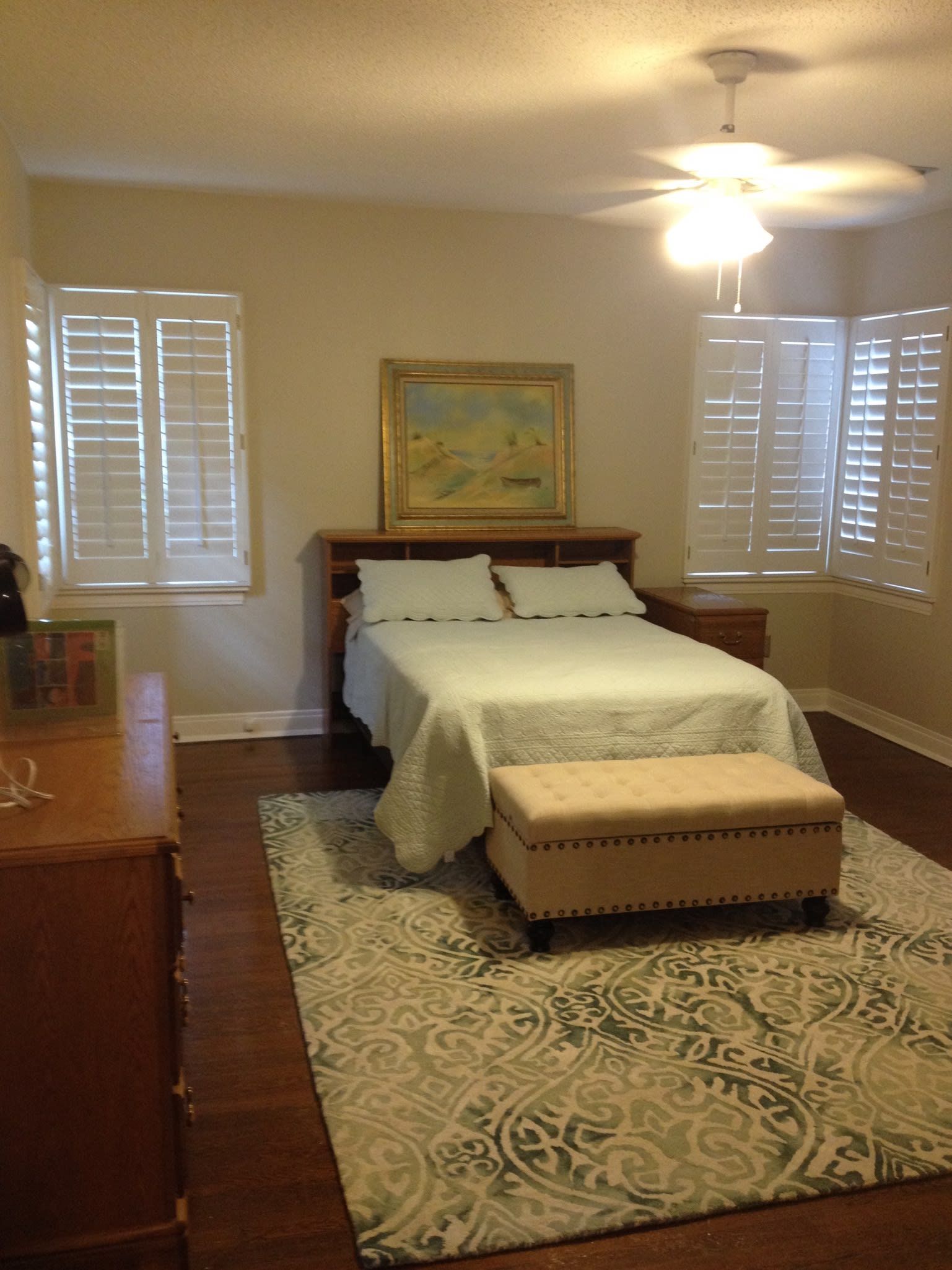 A bedroom with a bed , ottoman , dresser and shutters
