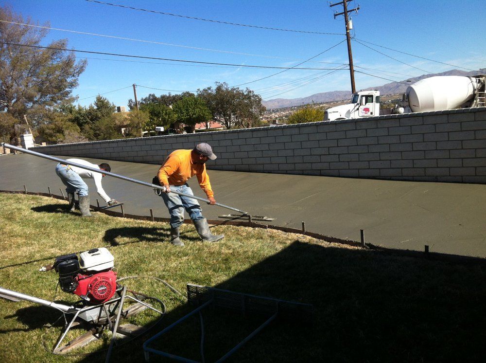 Two workers smoothing wet concrete with long tools outdoors; near a brick wall and yard.