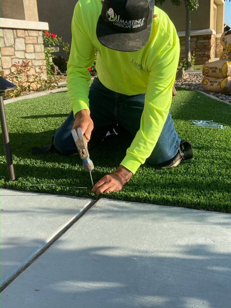 Man in neon shirt kneeling, hammering a metal strip into concrete near artificial turf.