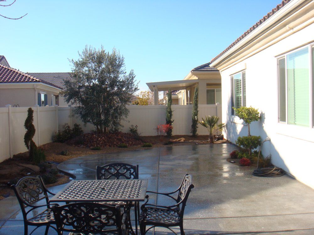 Patio with table and chairs, small trees, and a white fence under a blue sky.