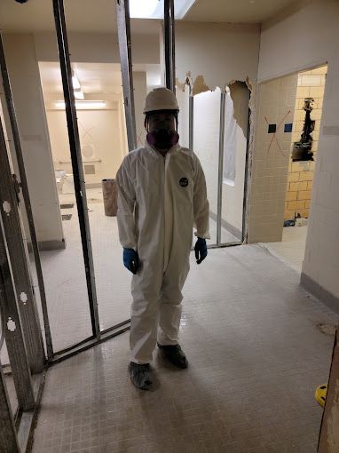 A man in a white coverall and hard hat is standing in a hallway.