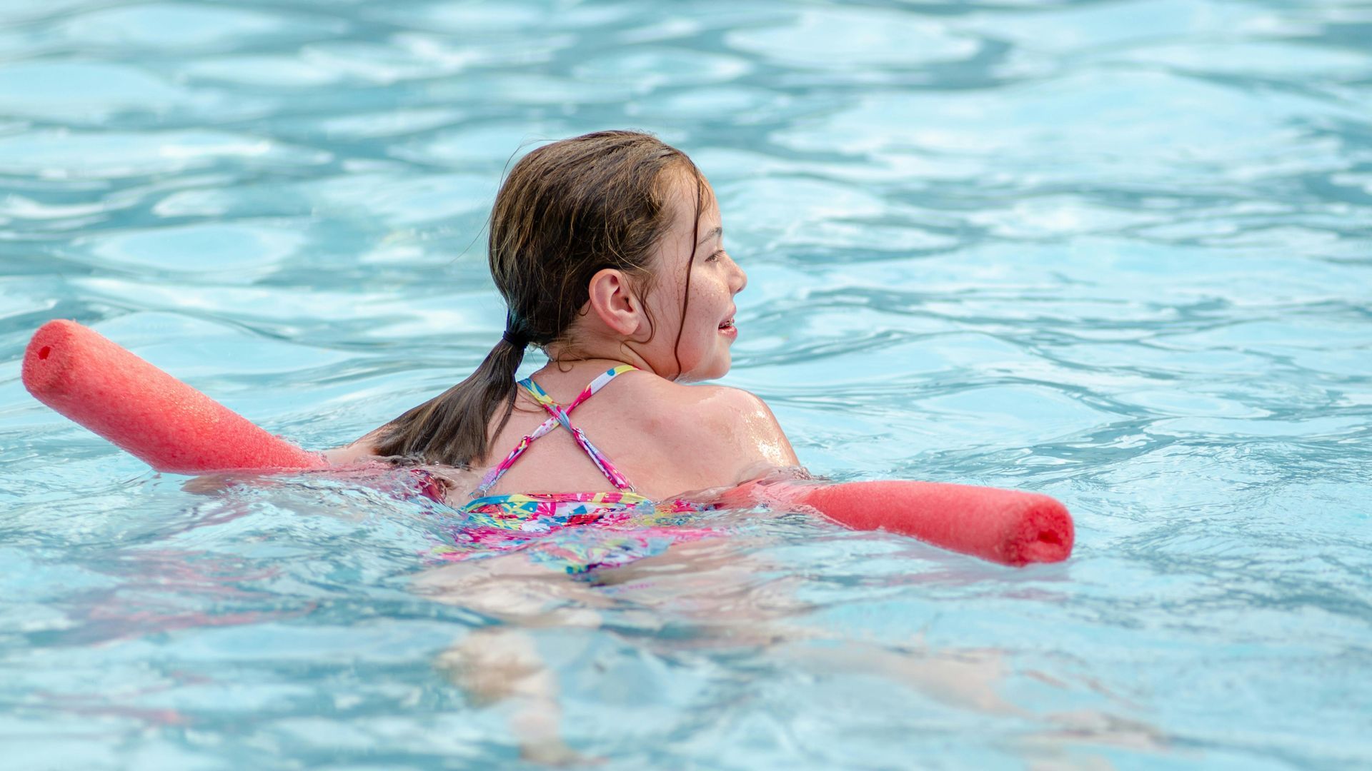 A young girl is swimming in a pool with a red noodle.