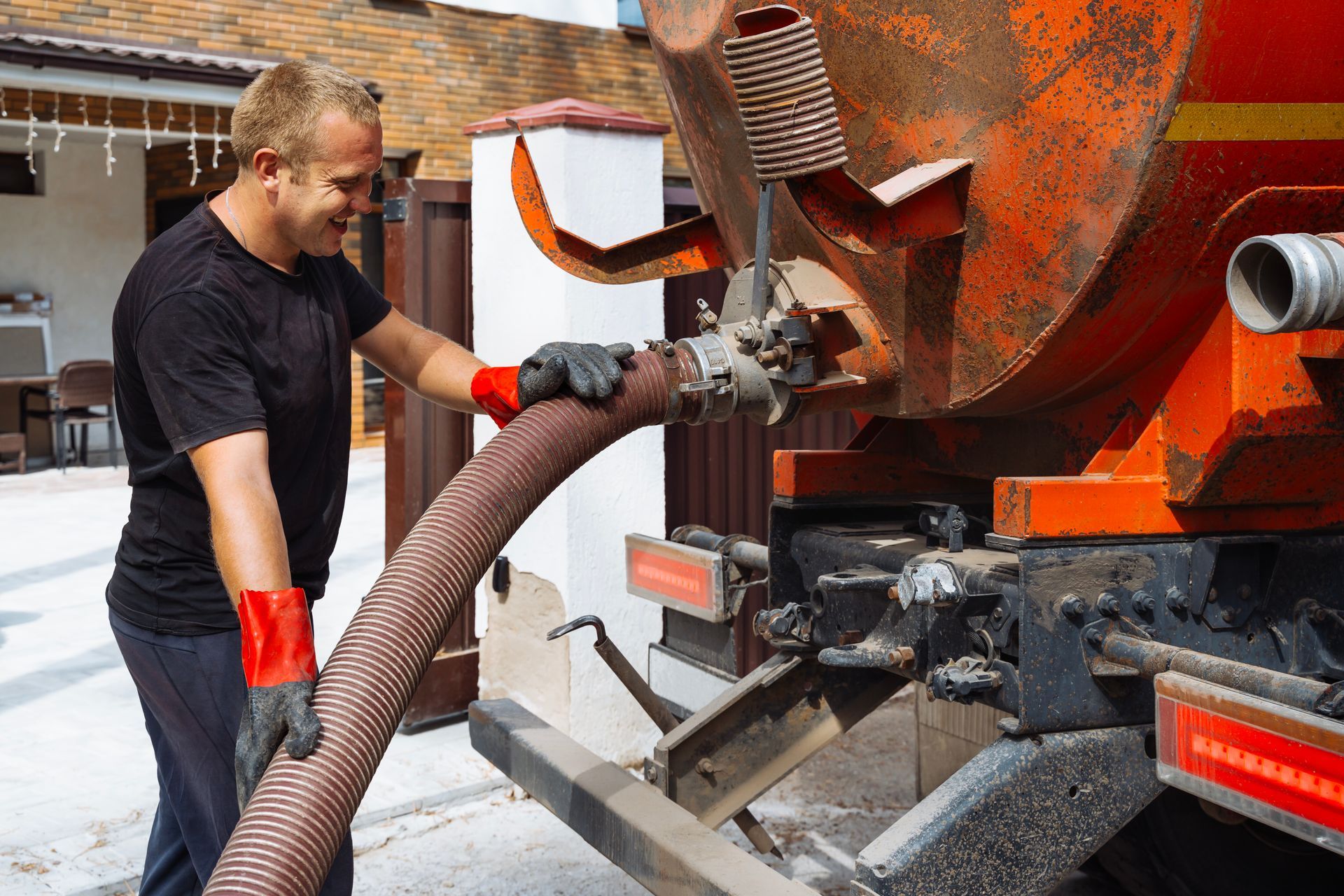 Man connecting hose to an orange septic truck; he is wearing gloves and smiling outdoors.
