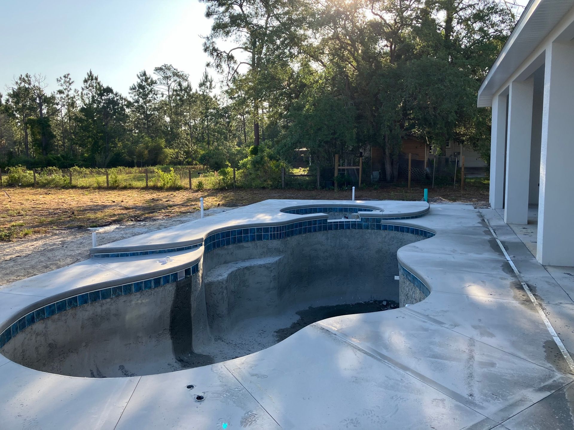 Empty pool under construction, surrounded by concrete, near a house and trees.