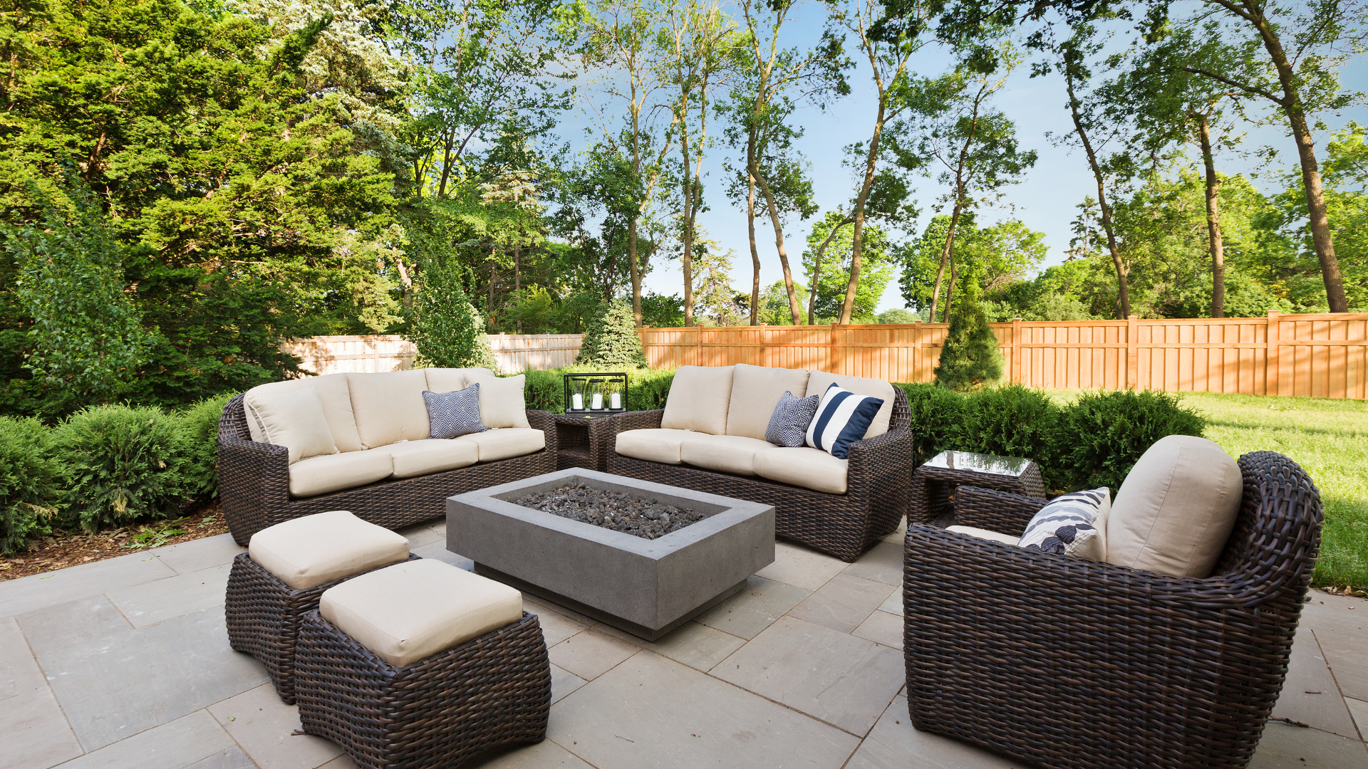 Outdoor patio with wicker furniture, including a fire pit, sofas, and seating. Surrounded by trees and bushes.