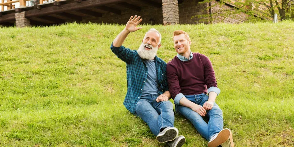 Two men are sitting on the grass waving at the camera.