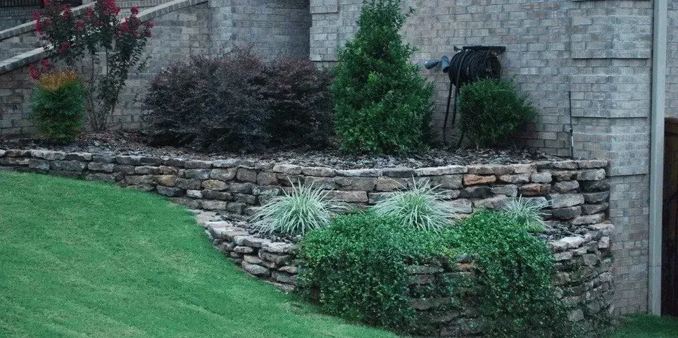 A stone wall surrounds a lush green lawn in front of a brick house.
