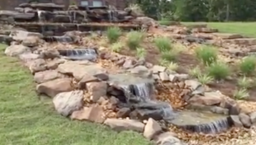 A waterfall is surrounded by rocks and grass in a yard.