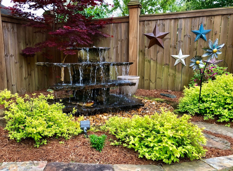 A water fountain is surrounded by plants and a wooden fence.