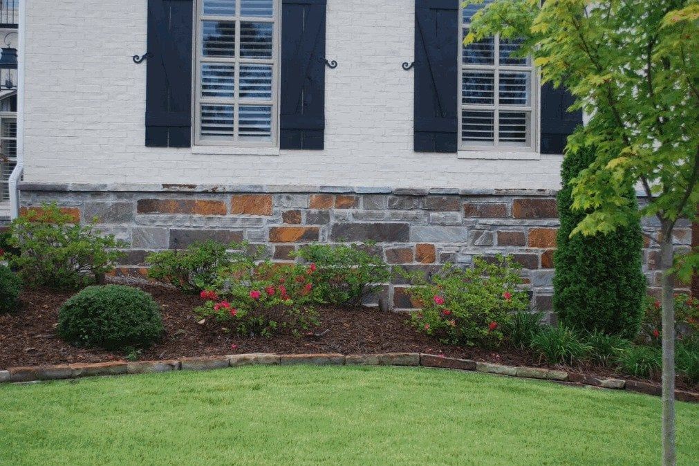 A white house with black shutters and a stone wall has a lush green lawn in front of it.