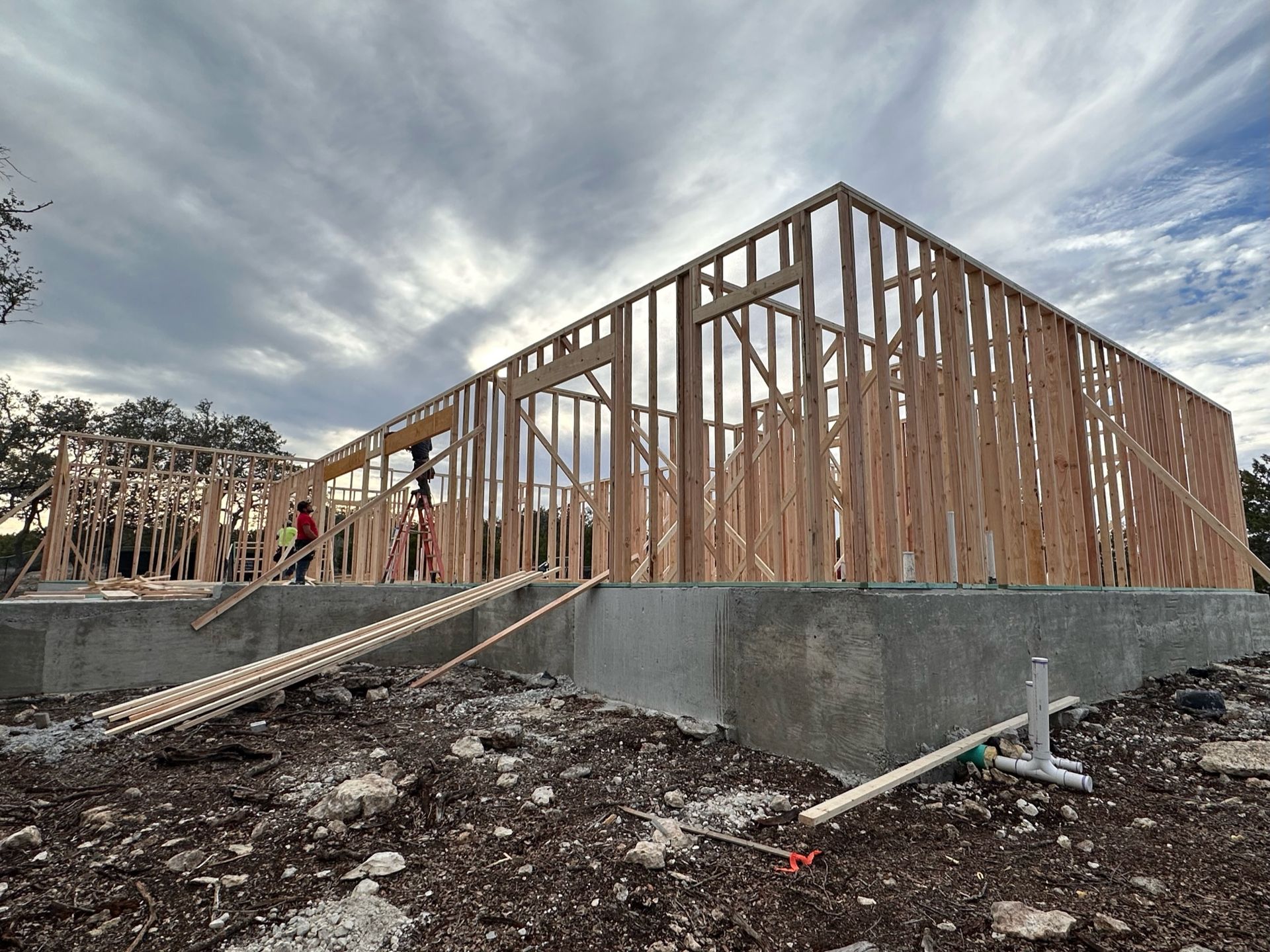 Wood-frame building under construction on a concrete foundation, with workers and a cloudy sky.