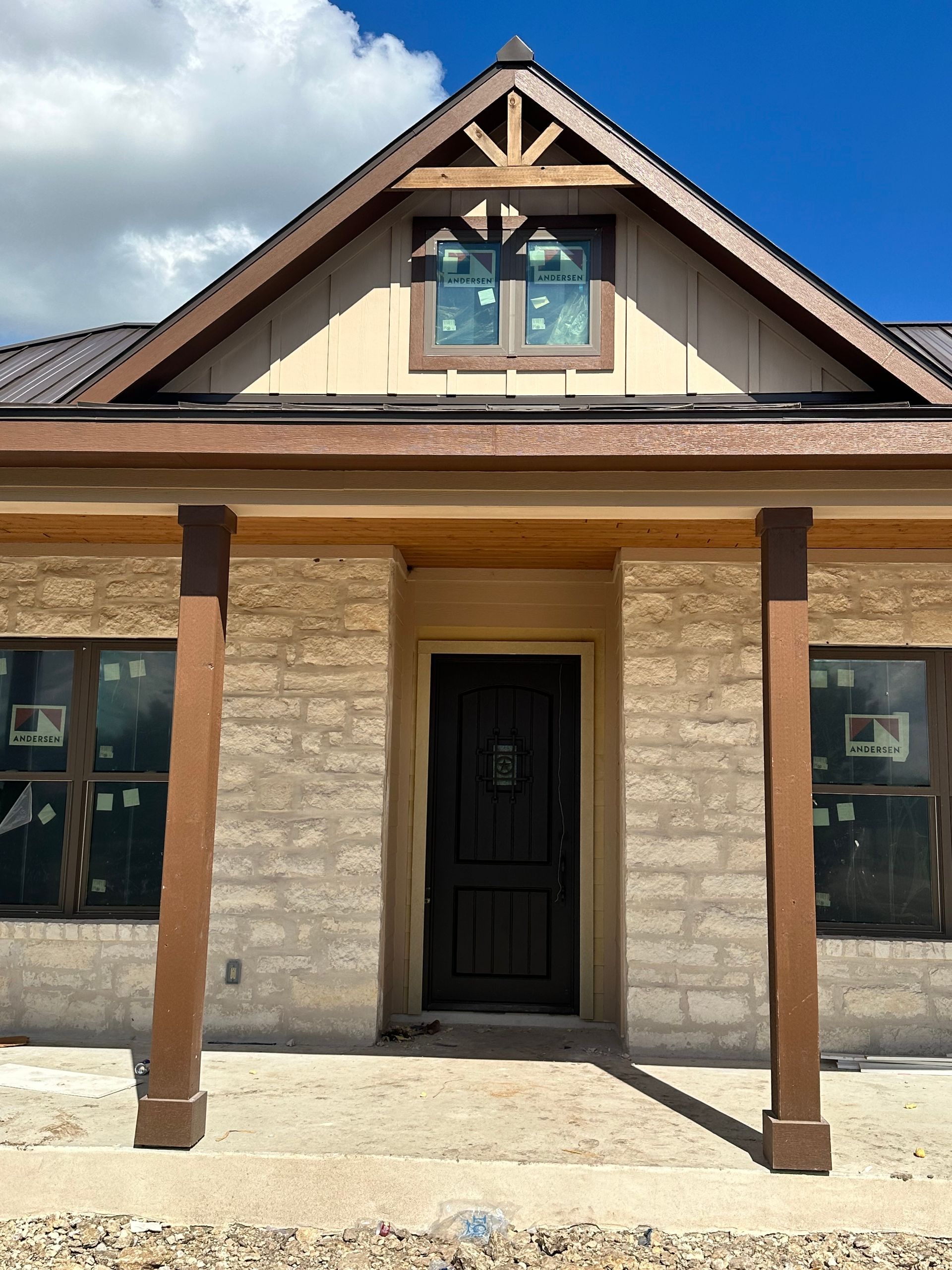 Front view of a tan stone house with a black door, wood posts, and a gabled roof under a blue sky