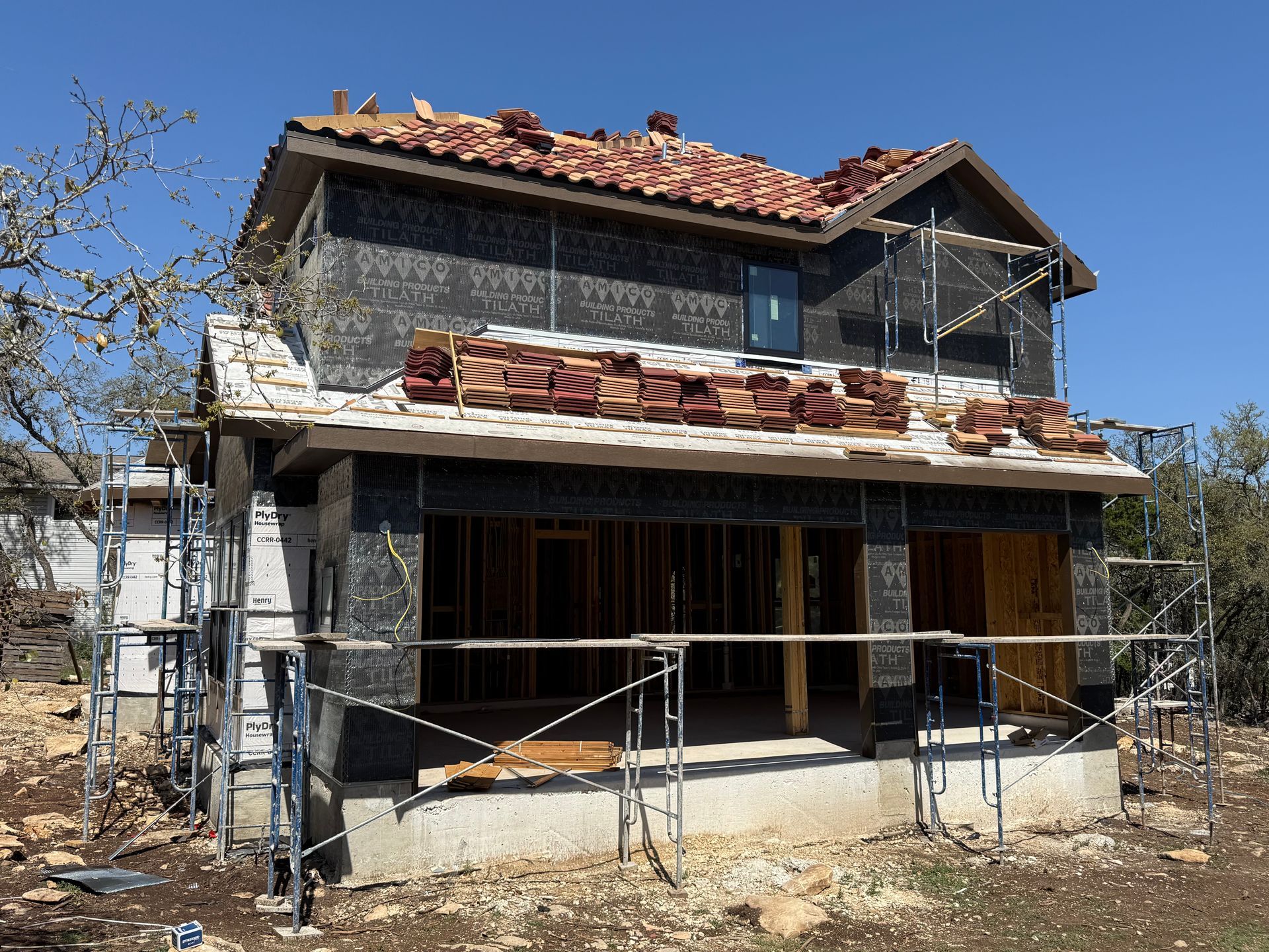 Partially renovated two-story house with scaffolding, exposed siding, and a red tile roof under a clear sky