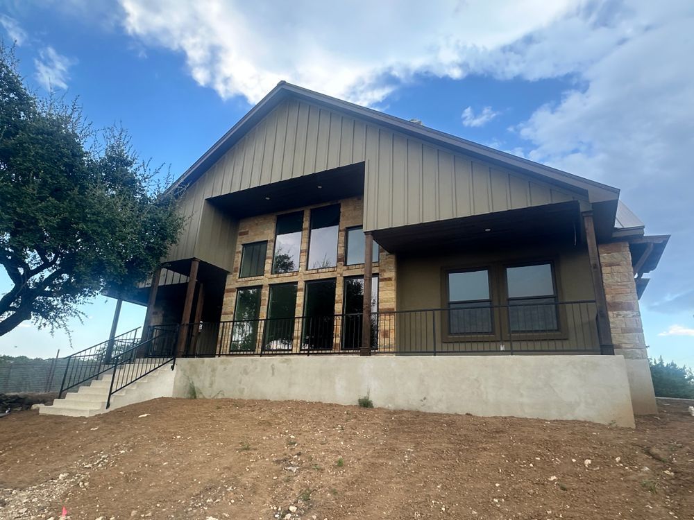 Modern house with tan siding, large windows, and a concrete porch on a dirt slope under a blue sky