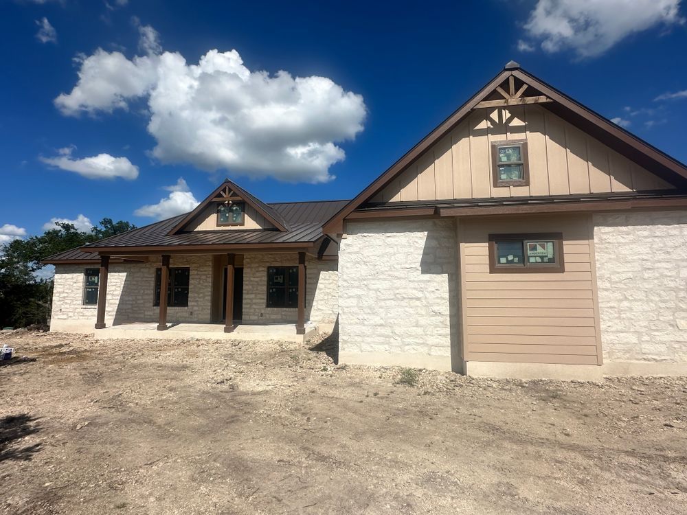 Stone house under construction with tan siding, brown trim, and a dirt foreground under a blue sky.