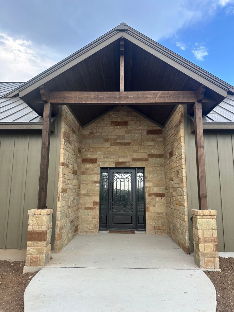Covered stone entryway with black double doors, wood beams, and concrete walkway under a gabled roof.