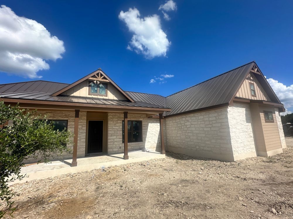 New single-story house with tan stone exterior and dark metal roof under a blue sky