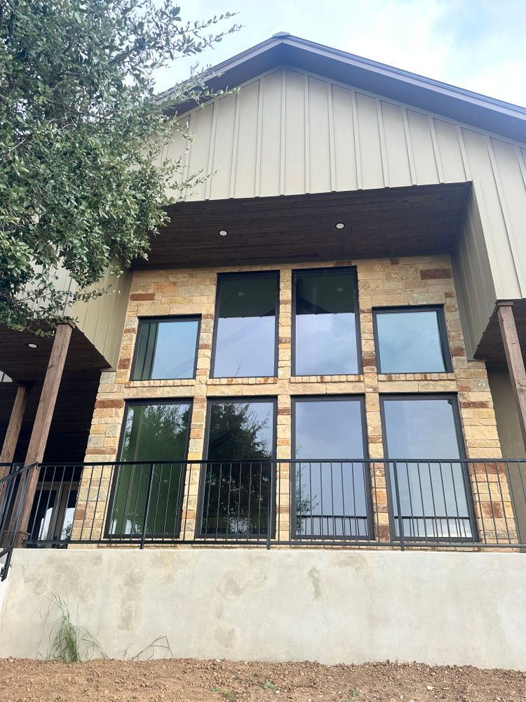 Modern house facade with large windows, stone columns, balcony railing, and trees on the left